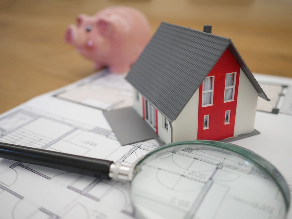 magnifying glass, house figurine and piggy bank rest on top of blueprints
